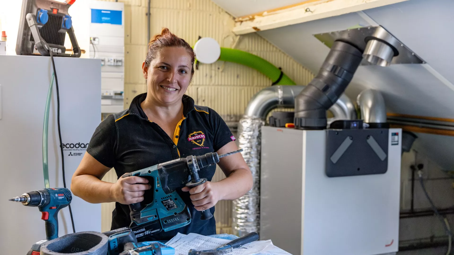 Werkvrouw staat met boormachine in de hand en grote glimlach op een zolder een klus te doen.
