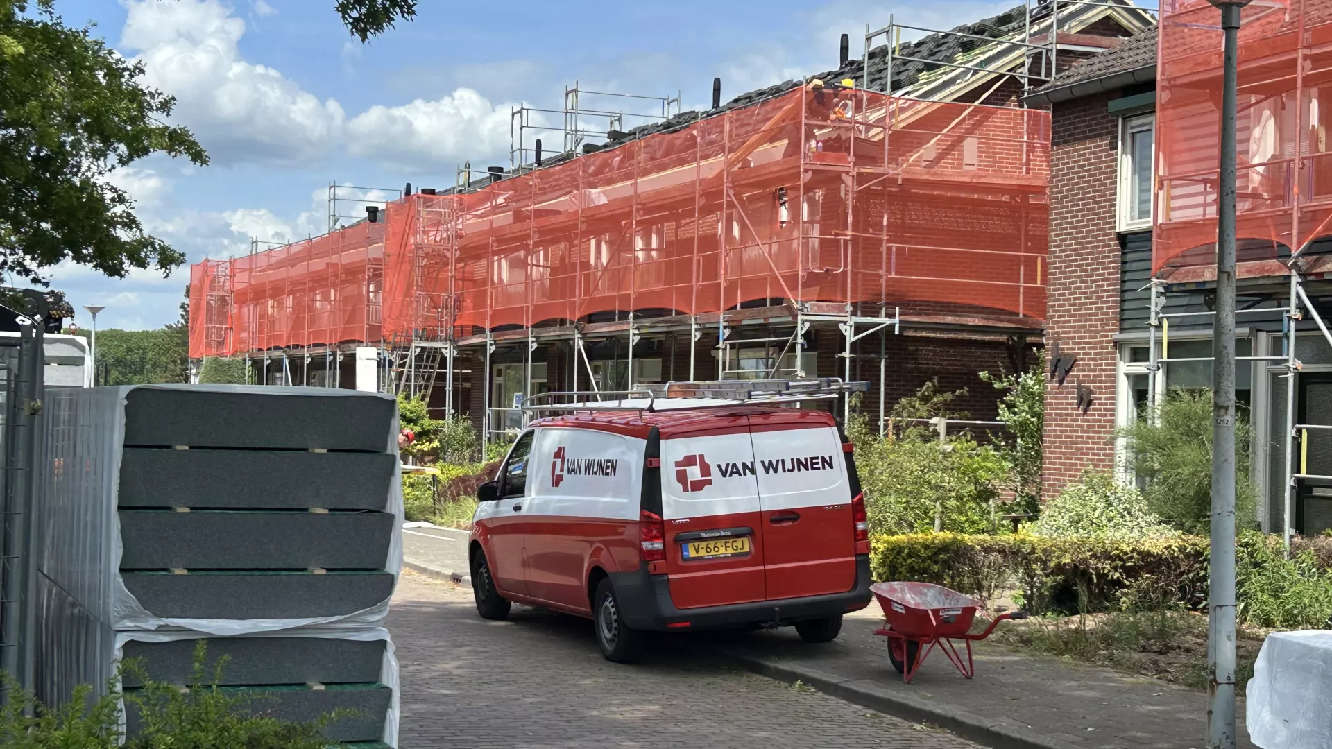 Rij met woningen die in de steigers staan. Om de steigers zit rood doorzichtig gaas. Voor de woningen staat een rode bus van een aannemer en liggen bouwplaten opgestapeld