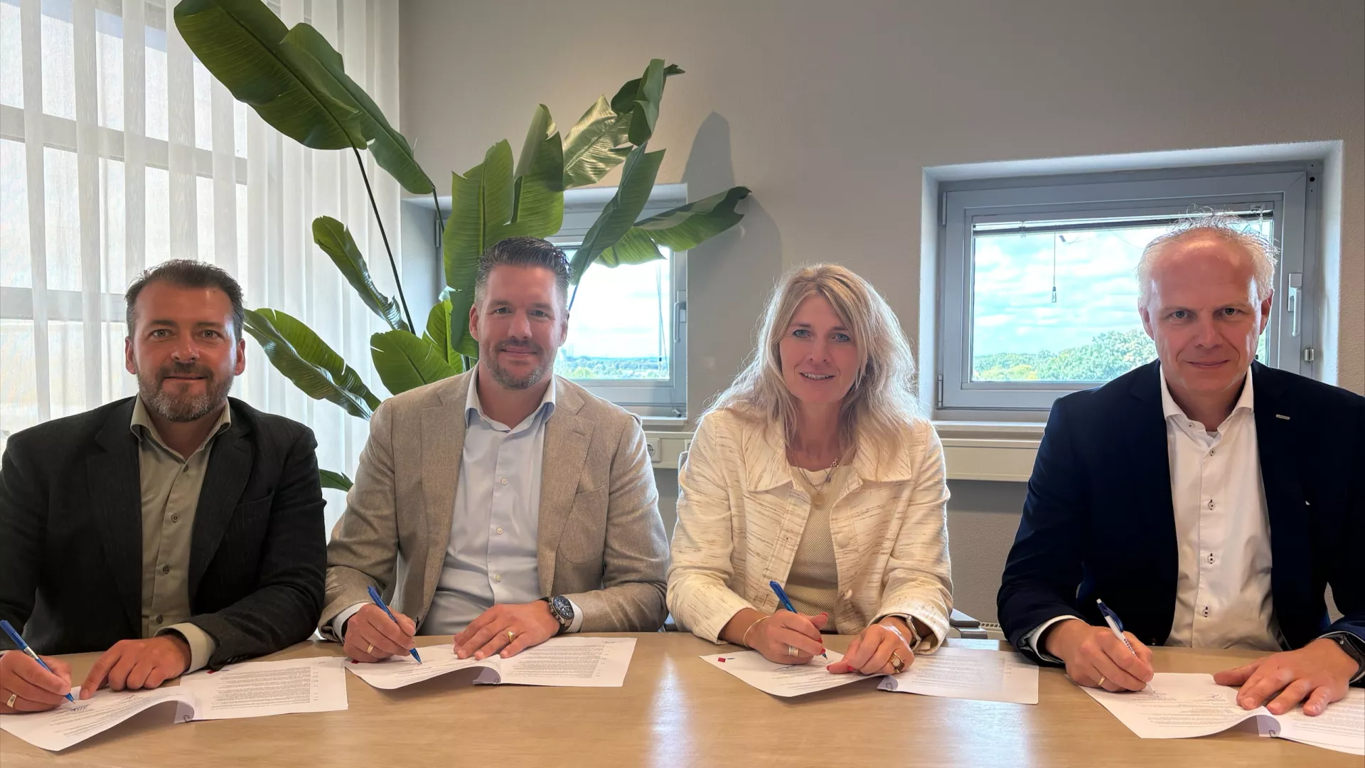 Drie mannen en een vrouw zitten aan tafel en zetten hun handtekening over een samenwerkingsovereenkomst