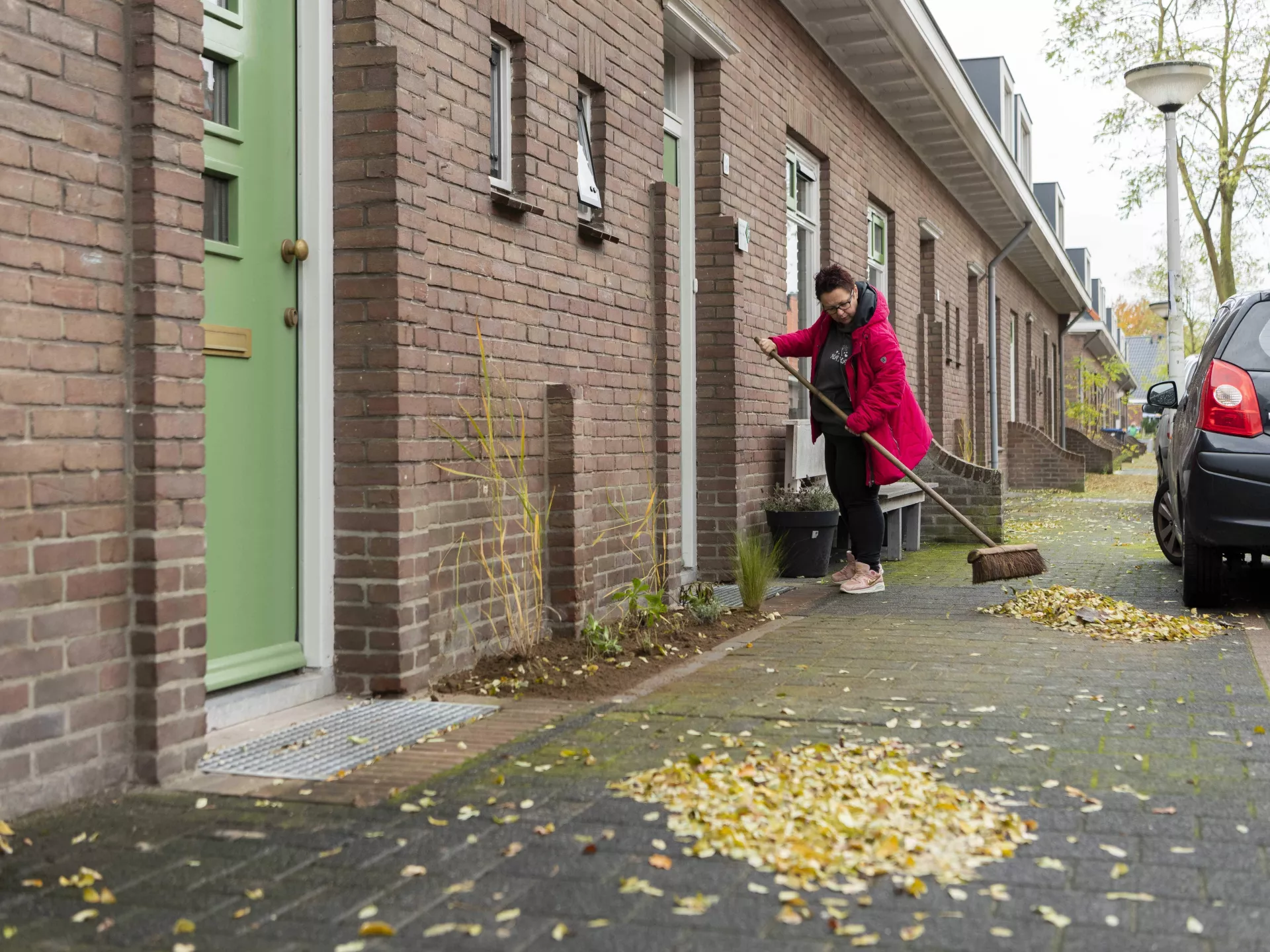 Vrouw met rode jas veegt voor haar huis de gevallen bladeren van de stoep