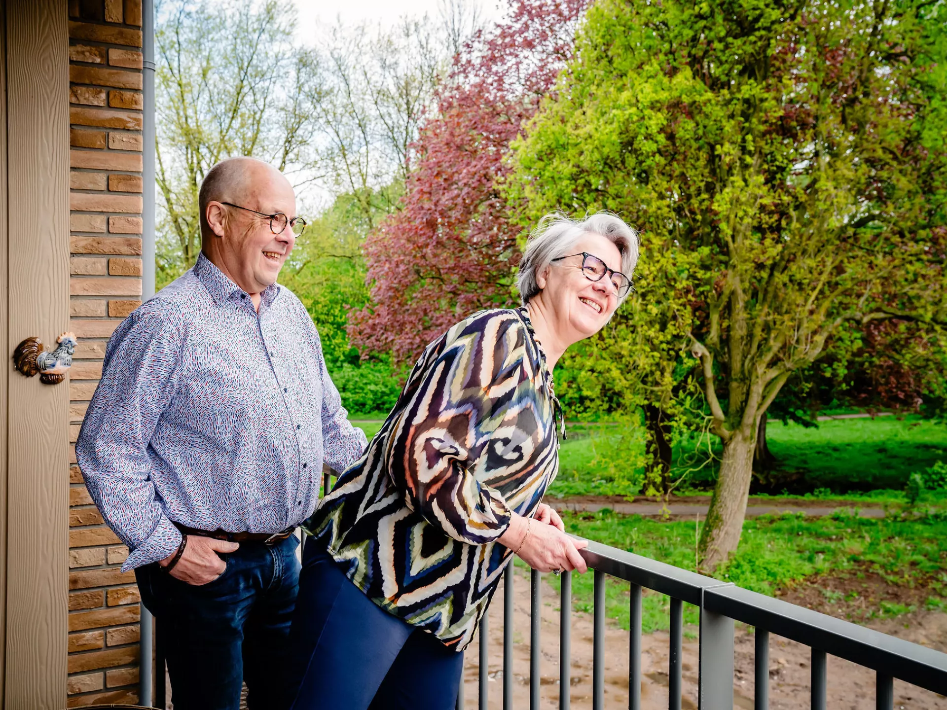 Oudere man en vrouw staan samen lachend op een balkon. Vrouw leunt over de balustrade