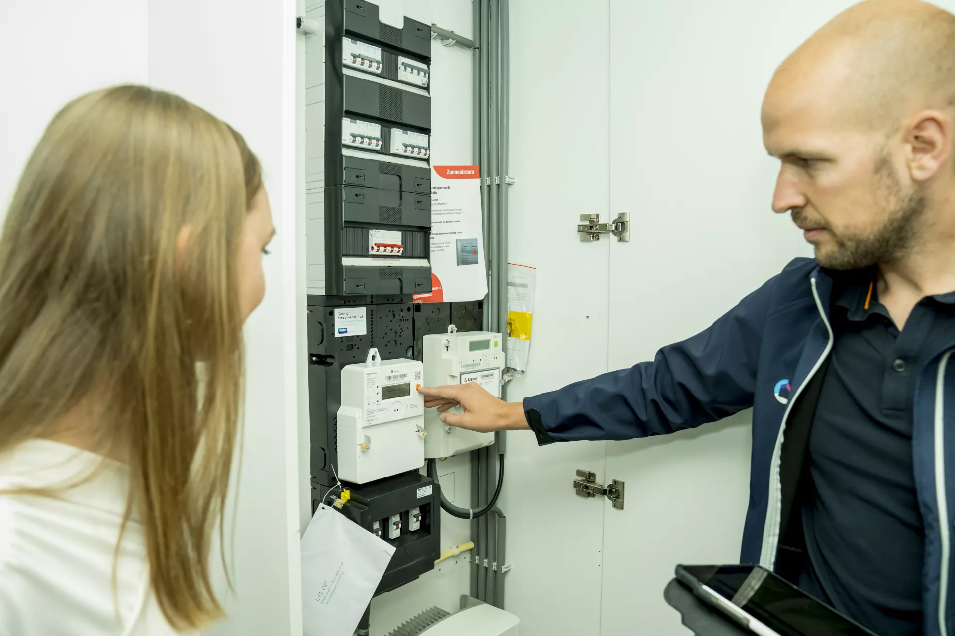 Man en vrouw staan bij een open meterkast. Man wijst naar een apparaatje in de meterkast