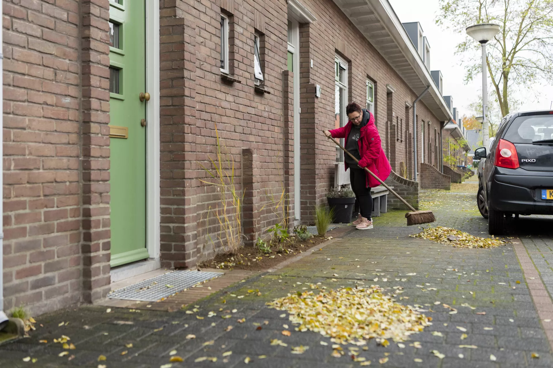 Vrouw met rode jas veegt voor haar huis de gevallen bladeren van de stoep