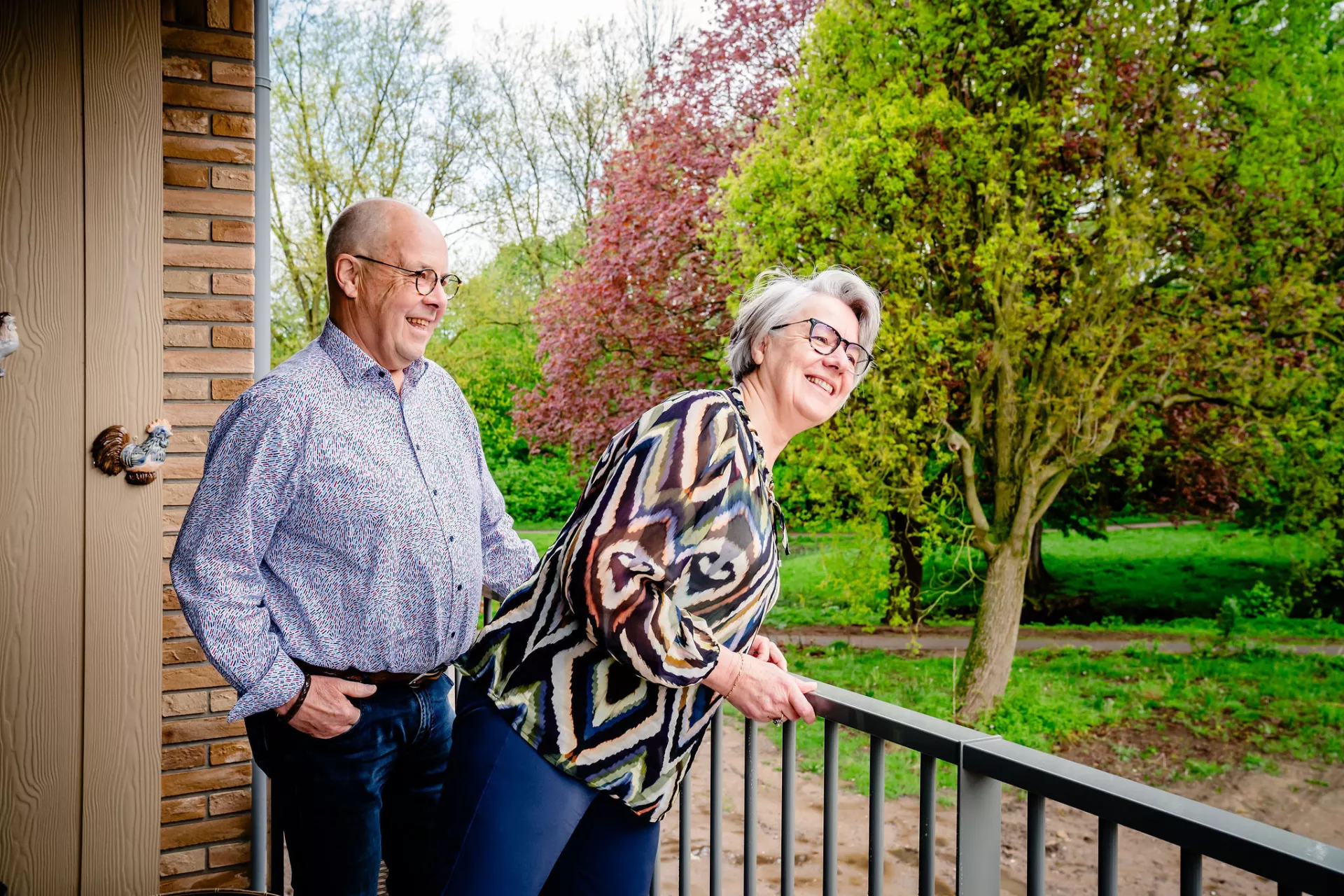 Oudere man en vrouw staan samen lachend op een balkon. Vrouw leunt over de balustrade