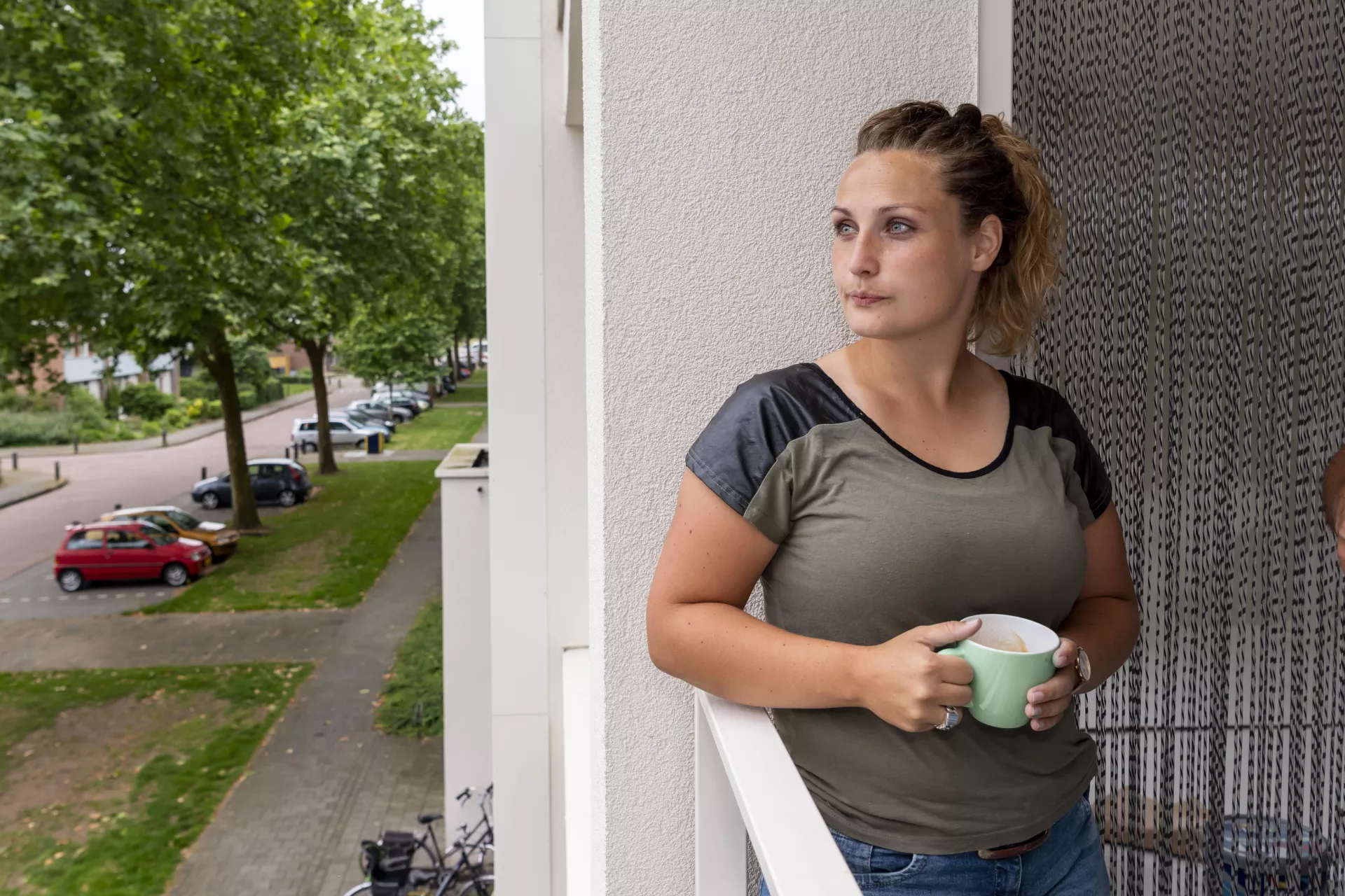 Dame staat met een kopje in haar hand op het balkon en staart voor zich uit