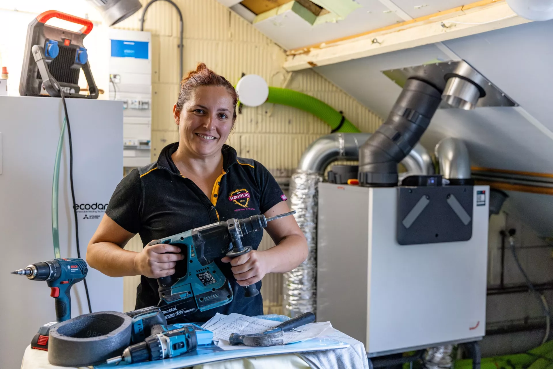 Werkvrouw staat met boormachine in de hand en grote glimlach op een zolder een klus te doen.