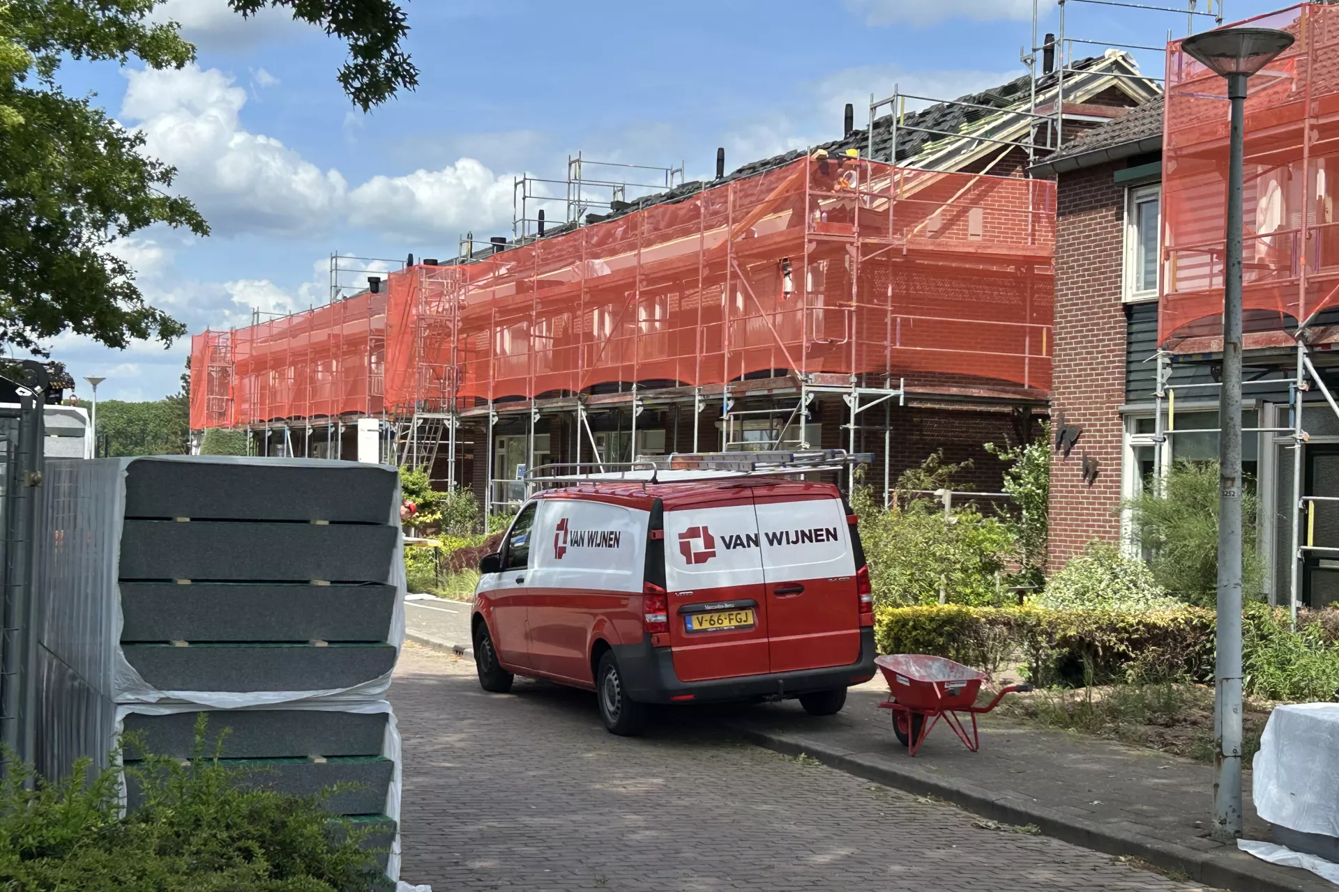Rij met woningen die in de steigers staan. Om de steigers zit rood doorzichtig gaas. Voor de woningen staat een rode bus van een aannemer en liggen bouwplaten opgestapeld