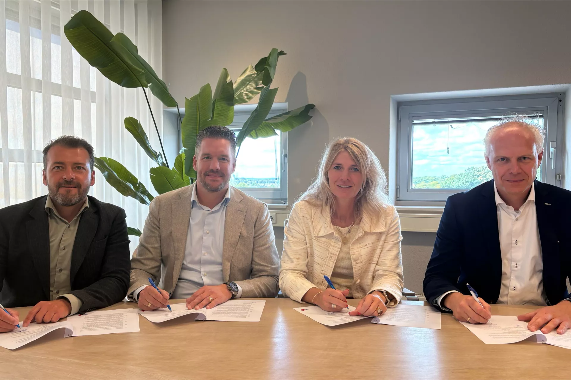 Drie mannen en een vrouw zitten aan tafel en zetten hun handtekening over een samenwerkingsovereenkomst